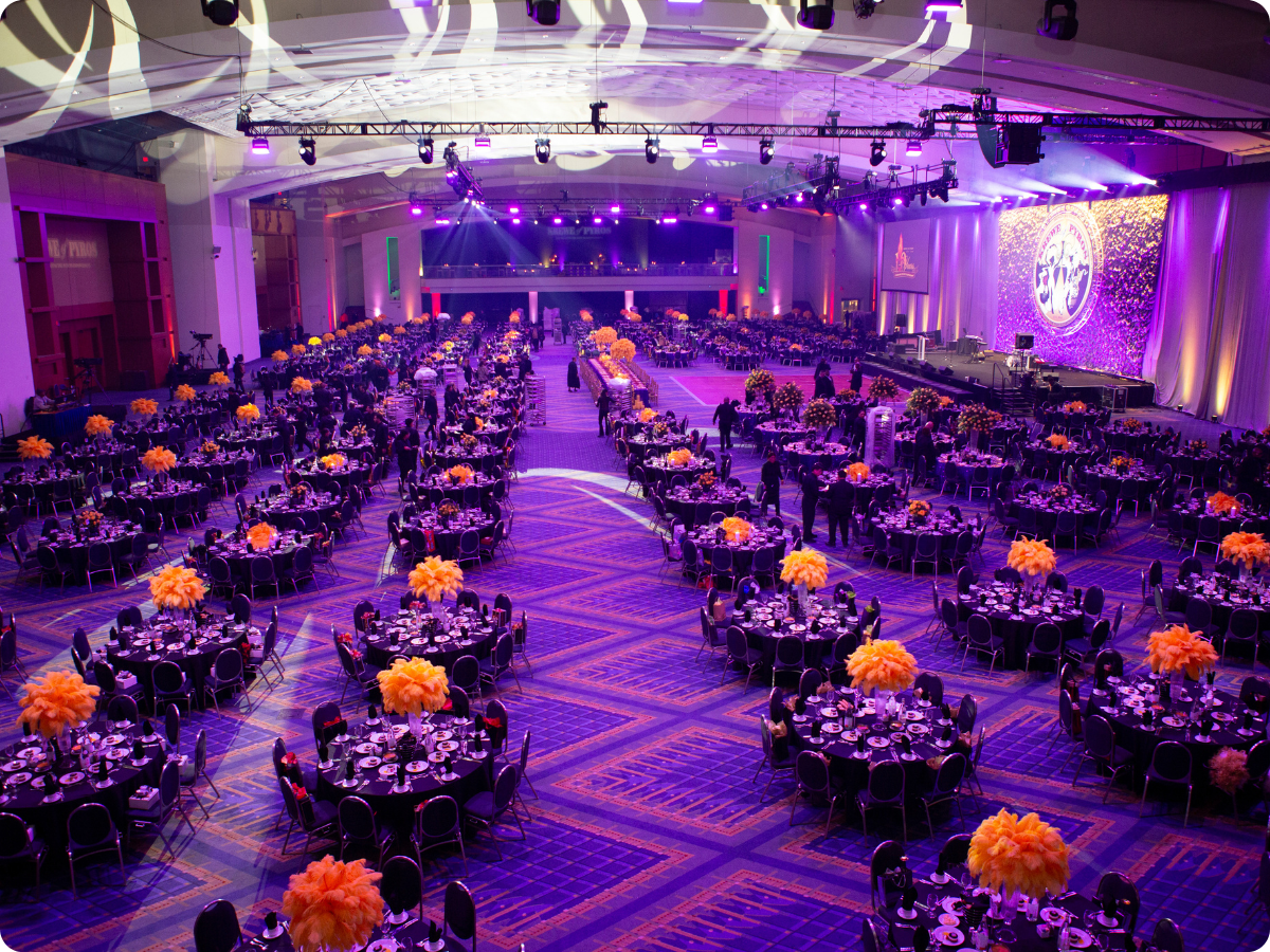 The Krewe of Pyros Annual Mardi Gras Ball ballroom at the Walter E. Washington Convention Center, set with hundreds of round tables and orange feather centerpieces under purple stage lighting